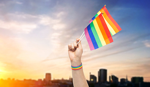 lgbt, same-sex relationships and homosexual concept - close up of male hand with gay pride awareness wristband holding rainbow flags over sunset in tallinn city background
