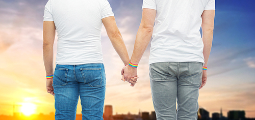 lgbt, same-sex relationships and homosexual concept - close up of male couple wearing gay pride rainbow awareness wristbands holding hands over sunset in tallinn city background