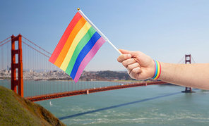 lgbt, same-sex relationships and homosexual concept - close up of male hand with gay pride awareness wristband holding rainbow flag over golden gate bridge in san francisco bay background