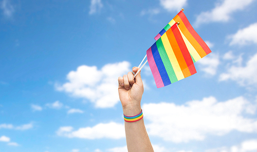 lgbt, same-sex relationships and homosexual concept - close up of male hand with gay pride awareness wristband holding  flags over blue sky and clouds background