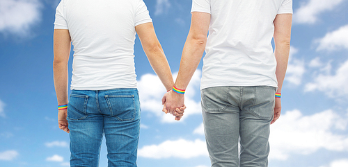 lgbt, same-sex relationships and homosexual concept - close up of male couple wearing gay pride rainbow awareness wristbands holding hands over blue sky and clouds background
