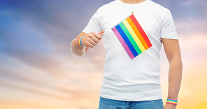 lgbt, same-sex relationships and homosexual concept - close up of man wearing gay pride awareness wristbands holding rainbow flag over evening sky background