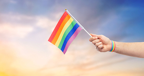 lgbt, same-sex relationships and homosexual concept - close up of male hand with gay pride awareness wristband holding rainbow flag over evening sky background
