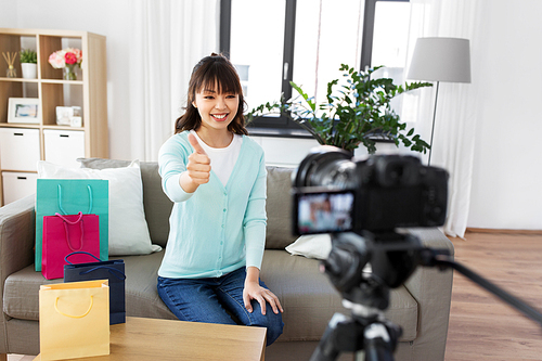 blogging, technology, videoblog and people concept - happy smiling asian woman or . with shopping bags recording video blog by camera and showing thumbs up gesture at home