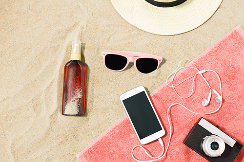 vacation and summer holidays concept - smartphone with earphones and film camera on towel, straw hat, sunglasses and bottle of sunscreen oil on beach sand