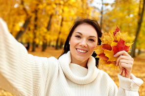 season and people concept - happy young woman with maple leaves taking selfie in autumn park