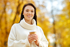 season, hot drinks and people concept - happy young woman drinking takeaway coffee or tea from disposable paper cup at autumn park