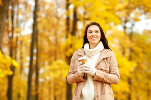 season, hot drinks and people concept - happy young woman drinking takeaway coffee or tea from disposable paper cup at autumn park