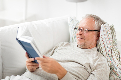 old age, leisure and people concept - senior man lying on sofa and reading book at home