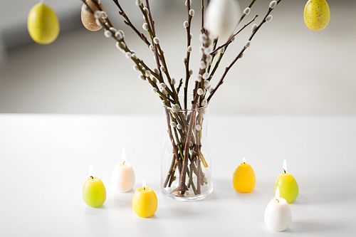 holidays and object concept - pussy willow branches decorated by easter eggs in vase and candles on table