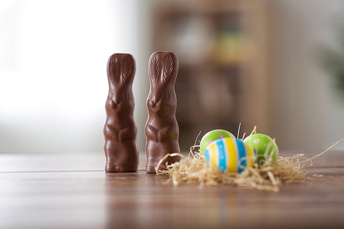 easter, holidays and object concept - close up of colored eggs in straw nest and chocolate bunnies on wooden table