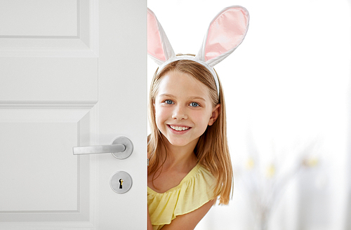 easter, holidays and people concept - happy girl wearing bunny ears peeking out door at home