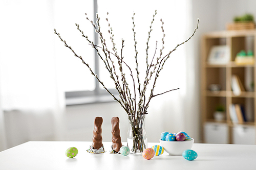 easter, holidays and object concept - pussy willow branches, colored eggs and chocolate bunnies on table at home