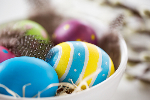easter, holidays, tradition and object concept - close up of colored eggs and quail feathers in bowl