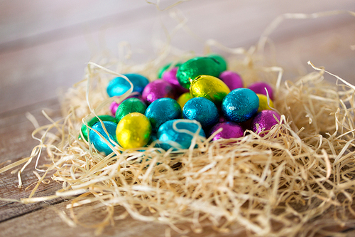 easter, confectionery and holidays concept - chocolate eggs in foil wrappers in straw nest on wooden table