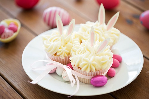 easter, food and holidays concept - frosted cupcakes with colored eggs and candies on wooden table