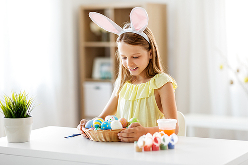 easter, holidays and people concept - happy girl wearing bunny ears headband with colored eggs at home