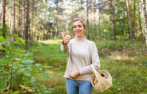 season and leisure people concept - young woman with mushrooms in wicker basket in forest showing thumbs up