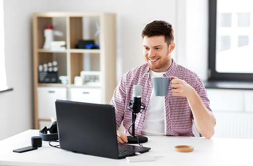 technology, mass media and podcast concept - happy male audio . with laptop computer and microphone drinking coffee and broadcasting at home office