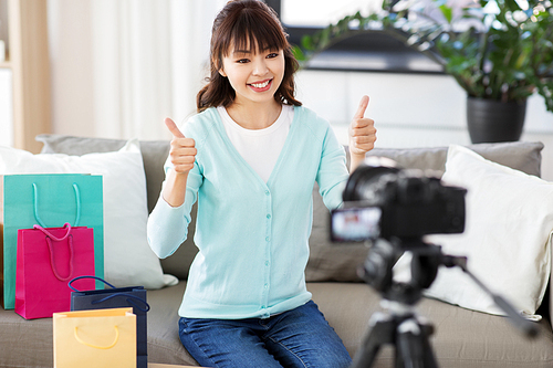 blogging, technology, videoblog and people concept - happy smiling asian woman or . with shopping bags recording video blog by camera and showing thumbs up gesture at home
