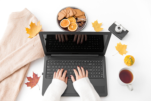 technology and season concept - woman's hands typing on laptop computer, cup of tea, film camera, autumn leaves and gingerbread cookies with sweater on white background