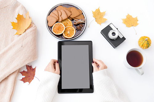 technology and season concept - woman's hands with tablet computer, cup of tea, film camera, autumn leaves and gingerbread cookies with dried orange slices on white background