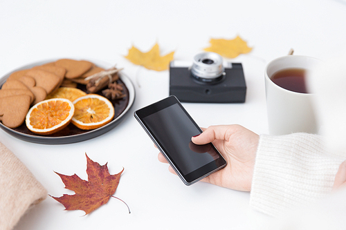 technology and season concept - woman's hand with smartphone, cup of tea, film camera, autumn leaves and gingerbread cookies with dried orange slices on white background