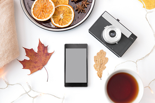 technology and season concept - smartphone, autumn leaves, film camera, dried orange slices with tea and garland lights on white background