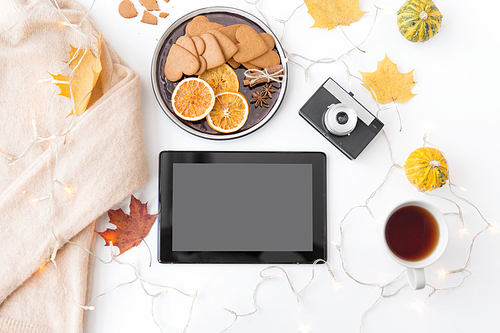 technology and season concept - tablet computer, film camera, autumn leaves, gingerbread cookies with dried orange slices and cup of tea on white background