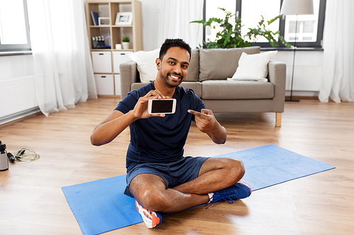 sport, technology and healthy lifestyle concept - smiling indian man with smartphone sitting on exercise mat at home