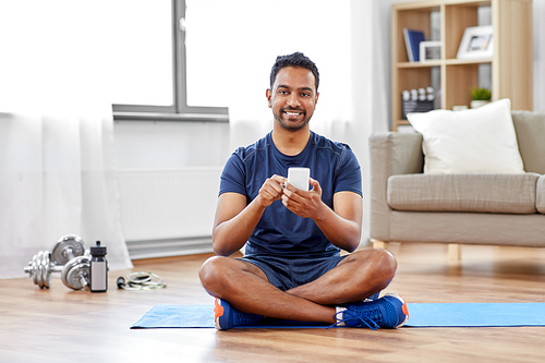 sport, technology and healthy lifestyle concept - smiling indian man with smartphone sitting on exercise mat at home