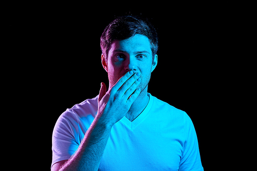 emotion, expression and people concept - portrait of young man in t-shirt covering his mouth by hand over ultra violet neon lights in dark room