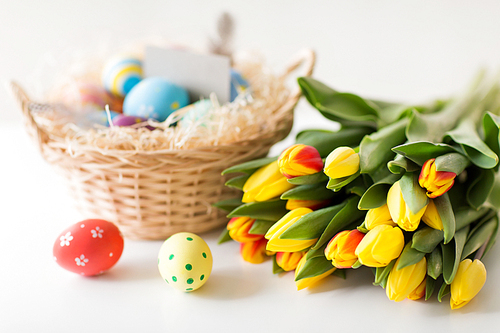 easter, holidays, tradition and object concept - close up of colored eggs in basket and tulip flowers on white background