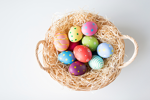 easter, holidays and tradition concept - colored eggs in basket with straw on white background