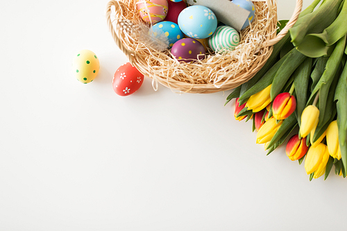 easter, holidays and tradition concept - close up of colored eggs in basket with straw and tulip flowers on white background