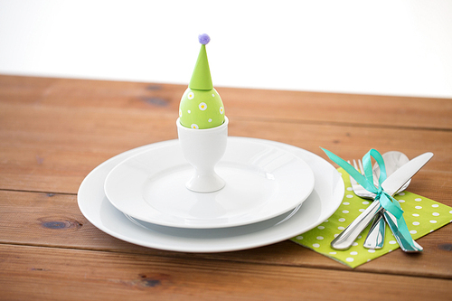 easter, holidays, tradition and object concept - green colored egg in cup holder, plates and cutlery on table at home