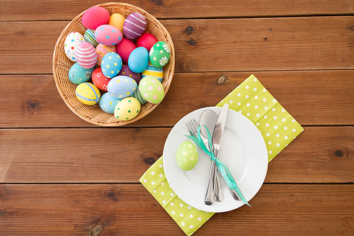 easter, holidays and object concept - colored eggs in basket, plates, cutlery and flowers on wooden table