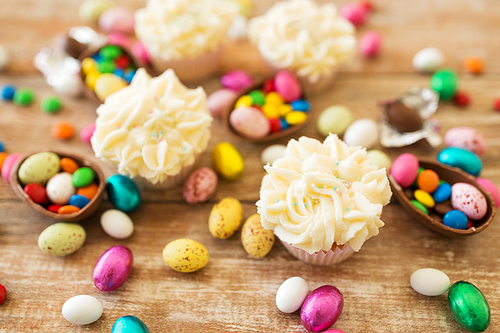 easter, food and holidays concept - close up of frosted cupcakes with chocolate eggs and candies on wooden table