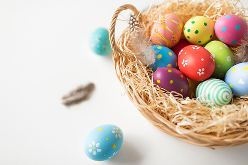 easter, holidays and tradition concept - close up of colored eggs in basket with straw and quail feathers on white background