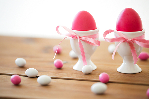 easter, food and holidays concept - pink colored eggs in ceramic cup holders with ribbon and candy drops on wooden table
