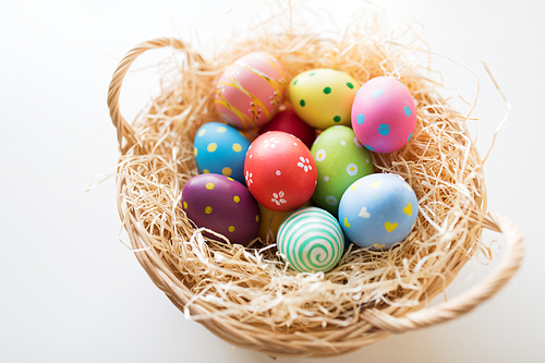easter, holidays and tradition concept - close up of colored eggs in basket with straw on white background