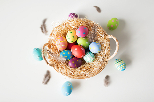 easter, holidays and tradition concept - colored eggs in basket with straw and quail feathers on white background