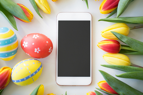 easter, holidays, tradition and object concept - smartphone with colored eggs and tulip flowers on white background