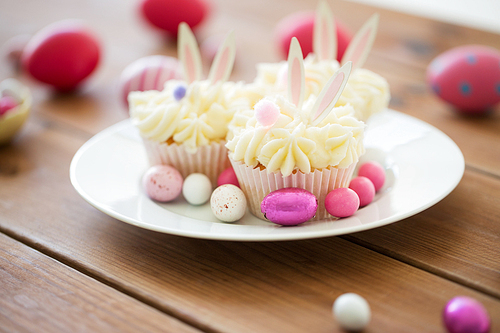 easter, food and holidays concept - frosted cupcakes with colored eggs and candies on wooden table