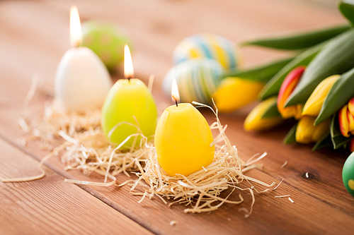 easter, holidays, tradition and object concept - three burning candles in shape of eggs and tulip flowers on wooden table