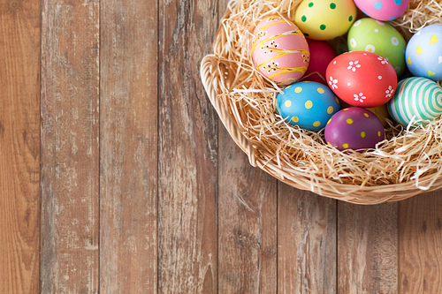 easter, holidays, tradition and object concept - close up of colored eggs in wicker basket over wooden boards background
