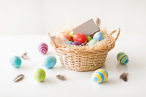 easter, holidays and object concept - colored eggs with greeting card in wicker basket on white background