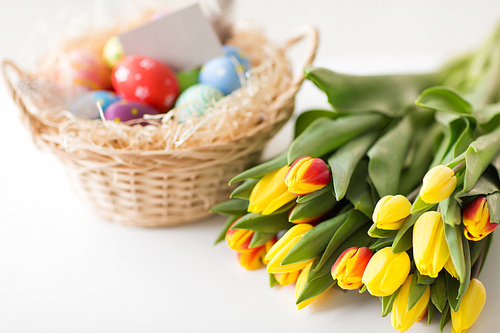 easter, holidays, tradition and object concept - close up of colored eggs in basket and tulip flowers on white background