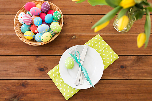 easter, holidays and object concept - colored eggs in basket, plates, cutlery and flowers on wooden table