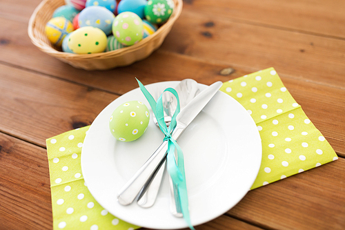 easter, holidays and table setting concept - close up of plate with cutlery and colored eggs on wooden table